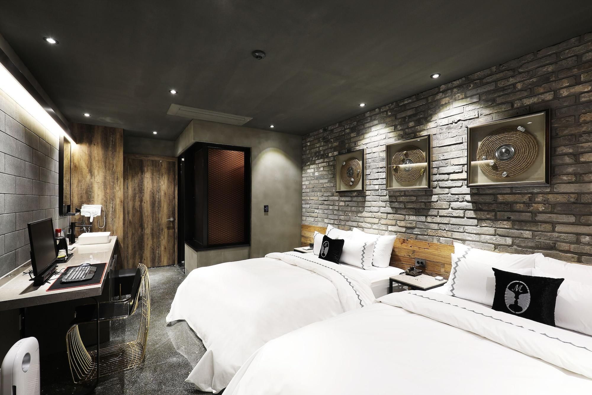 A hotel room with a brick wall, two beds, a desk with a computer, and a shower area.