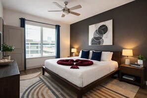 Ein Hotelzimmer mit Bett, zwei Nachttischen, einem Deckenventilator, einem Fenster mit Vorhängen, einem Wandbild im abstrakten Stil und Blick auf die Stadtlandschaft.