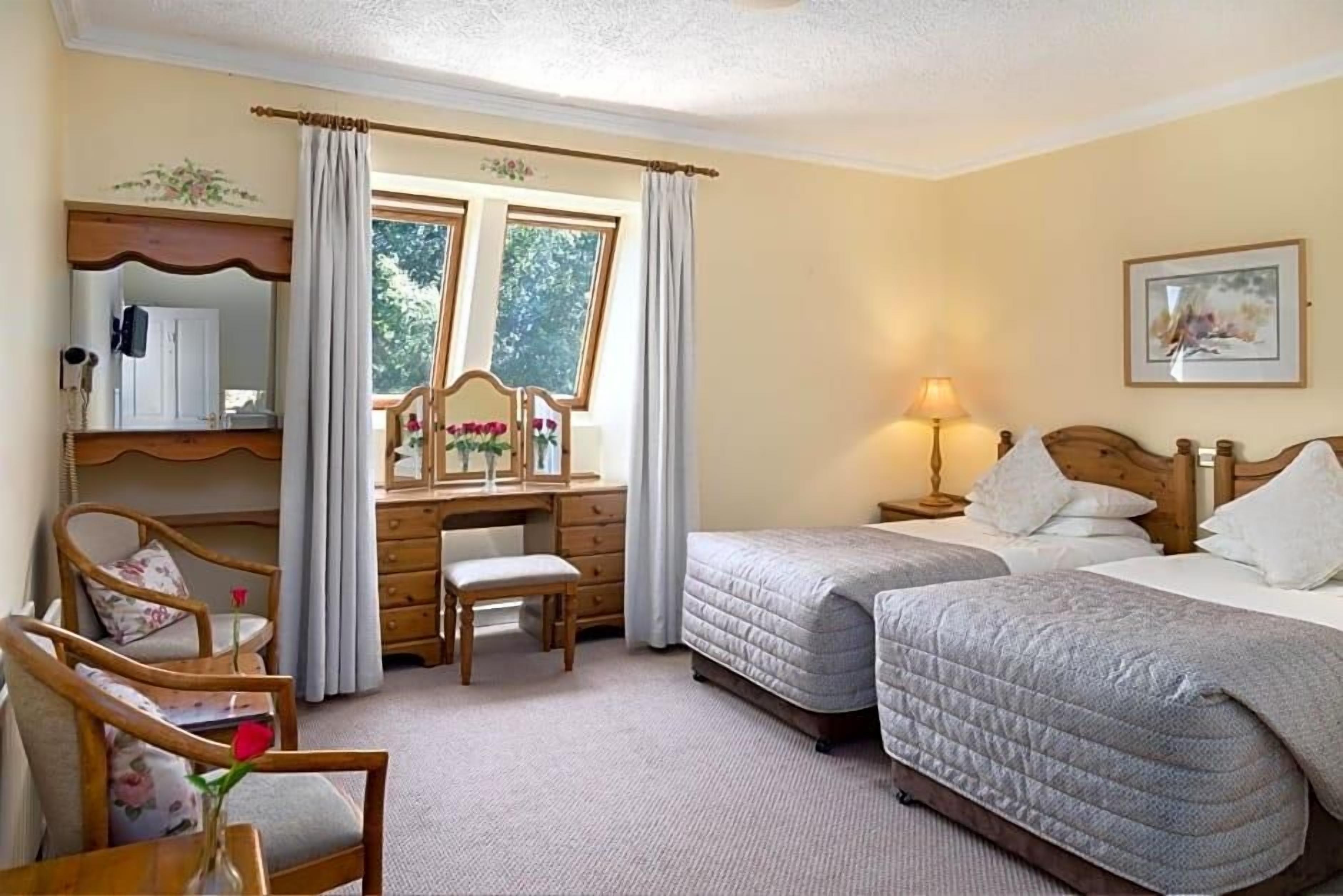 Un dormitorio con dos camas, un escritorio, una silla, una lámpara y una ventana con cortinas.