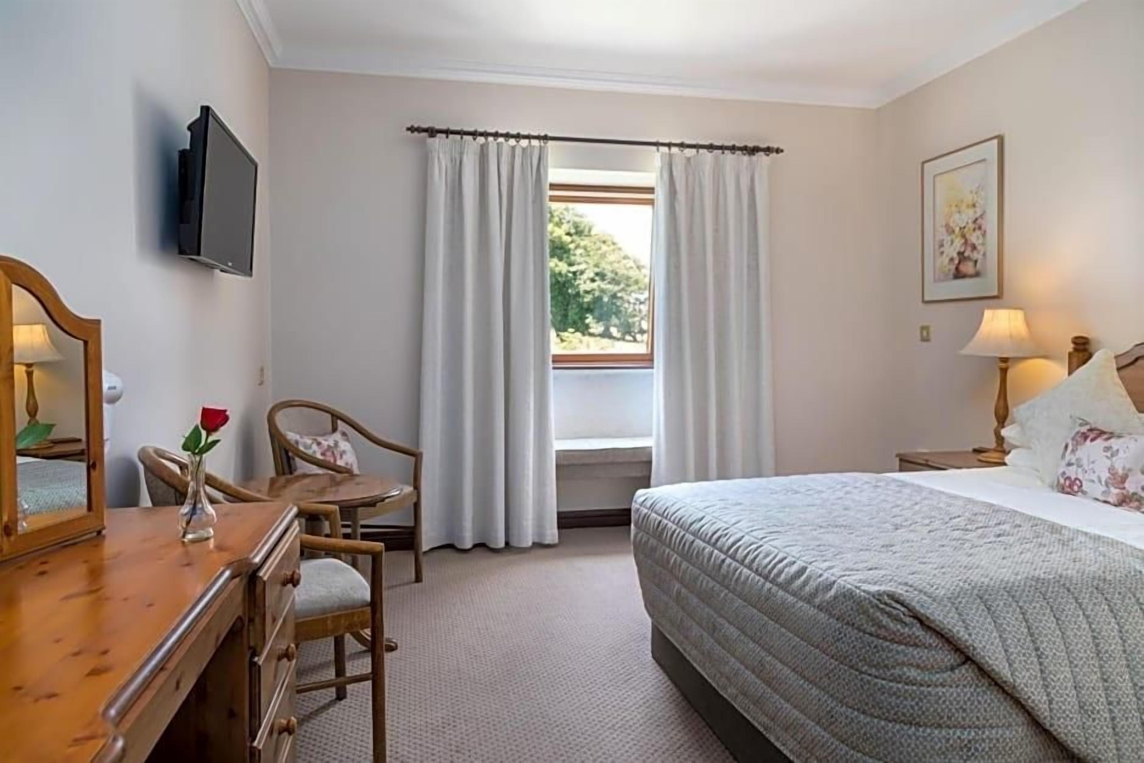 Habitación de hotel con cama, escritorio con silla, televisión, ventana con cortinas y un cuadro en la pared.