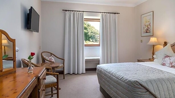 A hotel room with a bed, a desk with a chair, a television, a window with curtains, and a painting on the wall. - Kathleens Country (Killarney)