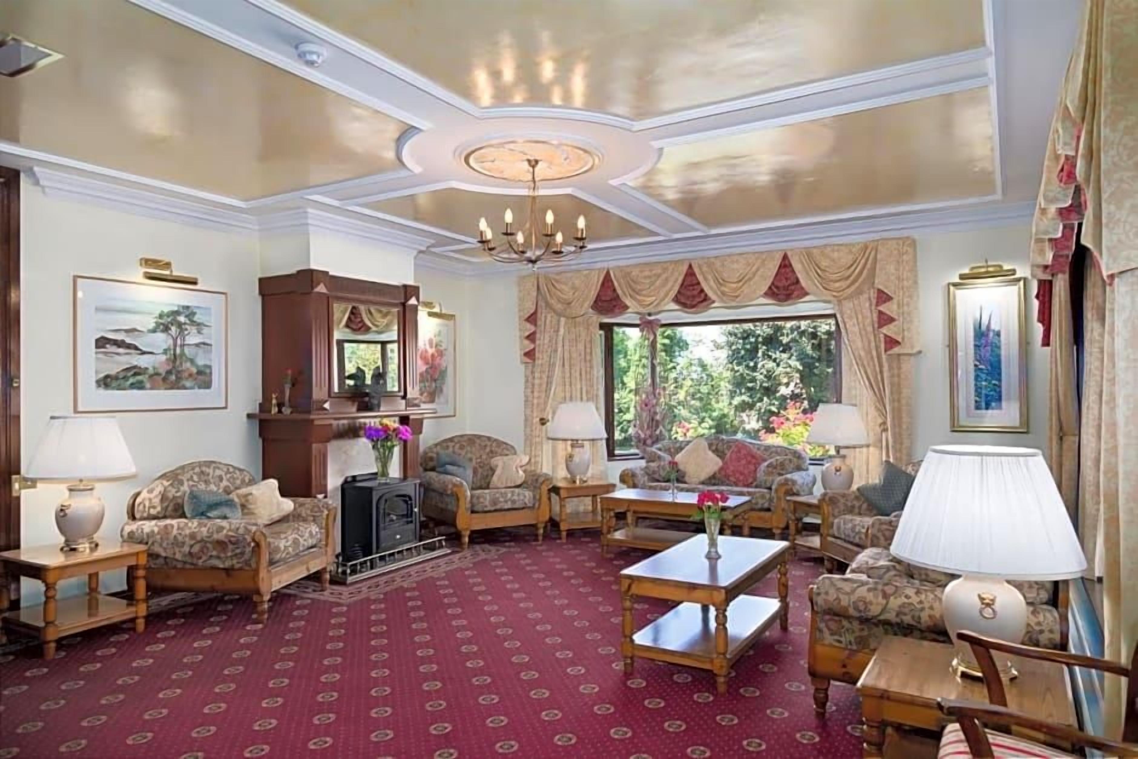 Una habitación con alfombra estampada, un candelabro y varios sofás y sillones dispuestos alrededor de una mesa de centro.