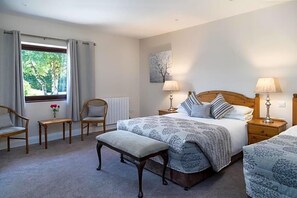 Un dormitorio con una cama grande, dos mesitas de noche con lámparas, un banco, dos sillas, una pequeña mesa con flores y una ventana con cortinas.