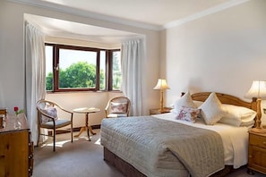 Habitación de hotel con cama, dos sillas, una mesita, una mesita de noche y ventana con cortinas.