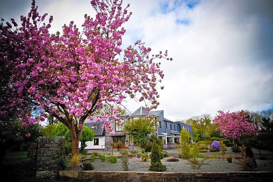 Una casa con pared de piedra, un jardín con diversas plantas y un gran árbol de flores rosas.