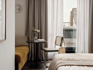 A hotel room with a bed, a small table with a vase, a chair, and a view of a building through the window. - Hotello Schwabing (Munich)