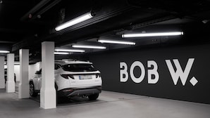 Eine Parkgarage mit einem weißen SUV, der in einem Platz mit der Nummer 22 geparkt ist. An der Wand sind große weiße Buchstaben zu sehen, die 'BOB W.' bilden.