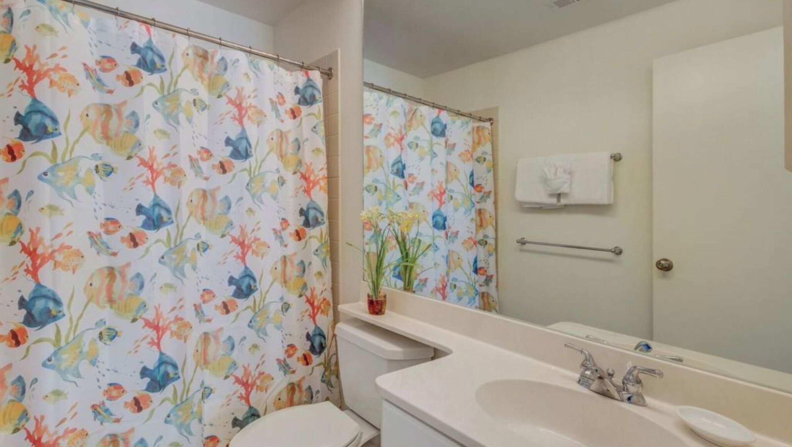 Ein Badezimmer mit Duschvorhang mit Fischmuster, Toilette, Waschbecken mit Spiegel und Handtuchhalter.