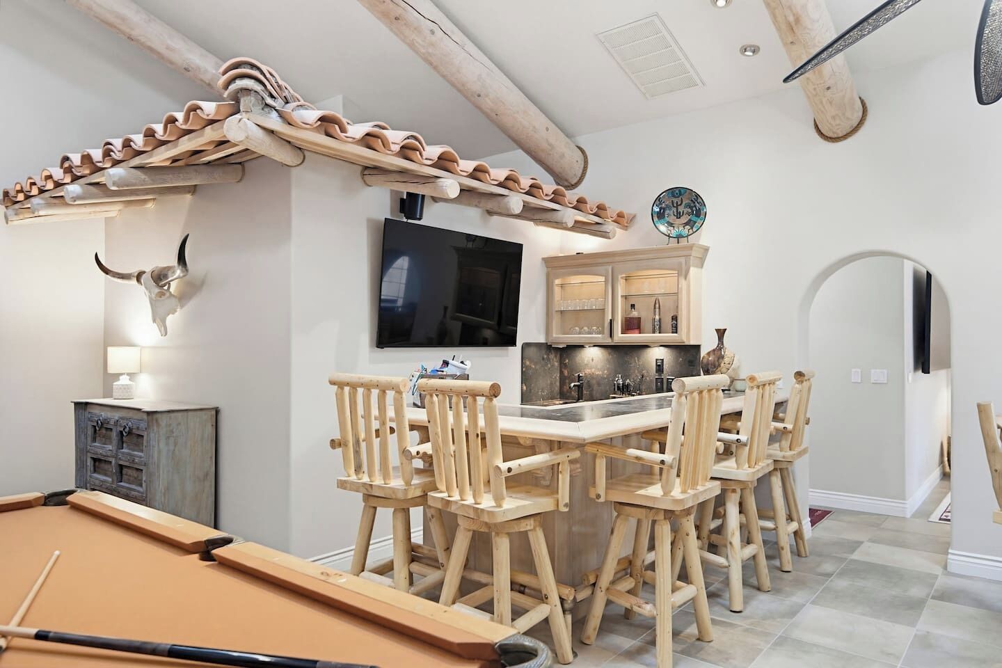 Une table de billard, des chaises en bois, un téléviseur, un coin cuisine et une décoration représentant un crâne de taureau.