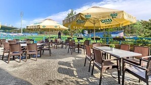 An outdoor seating area with tables and chairs, umbrellas, and a view of a sports field. - Land-gut-hotel Olympic (Wronki)