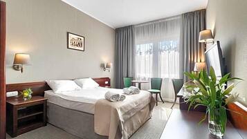 Et hotelværelse med seng, sengeborde, skrivebord, stol, lampe, et billede på væggen og en plante.