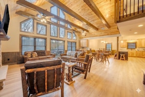 A spacious living area with wooden furniture, large windows, and a kitchen in the background.