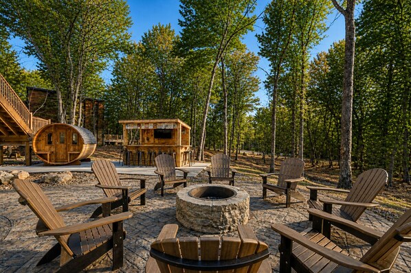 A rustic outdoor seating area with Adirondack chairs, a fire pit, and a wooden structure with a grill.