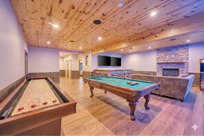 A pool table, a billiards table, a sofa, and a stone fireplace in a room with wooden ceilings and walls.
