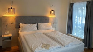 A neatly made bed with white linens, a headboard with a geometric pattern, bedside lamps, and a window with curtains. - Stadt-gut-hotel Astra (Ulm)