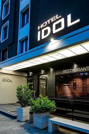 En hotelindgang med et skilt, der siger 'HOTEL IDOL', et receptionområde og en restaurant, der kan ses gennem glasset.