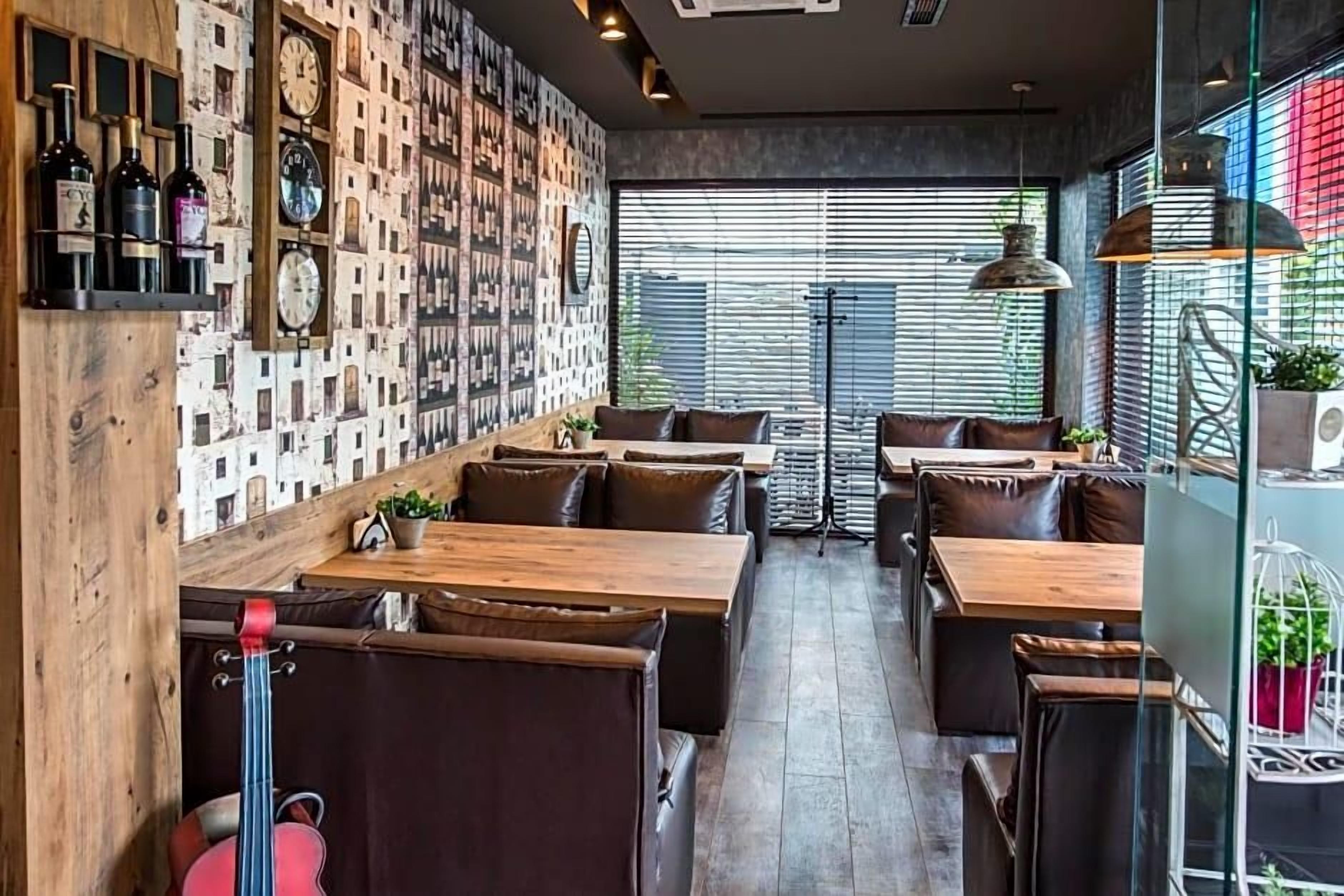 Interior de restaurante com mesas e cadeiras de madeira, bancos acolchoados, uma parede com relógios e grandes janelas.