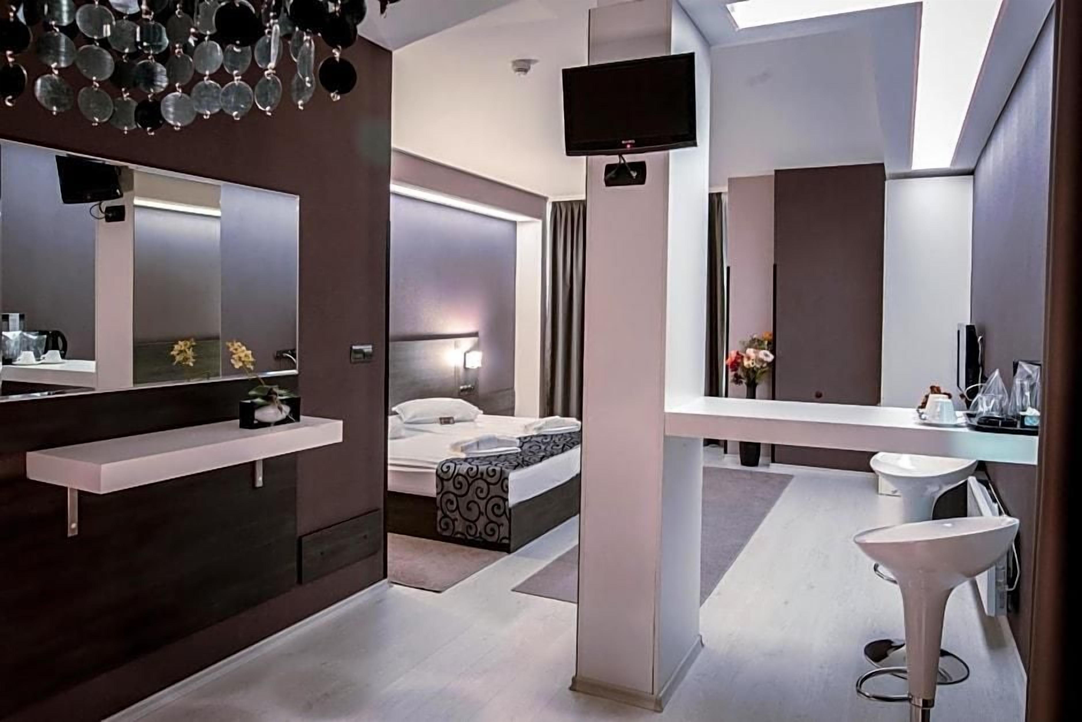Habitación de hotel moderna con cama, televisor, espejo grande, tocador con lavamanos y un taburete pequeño.