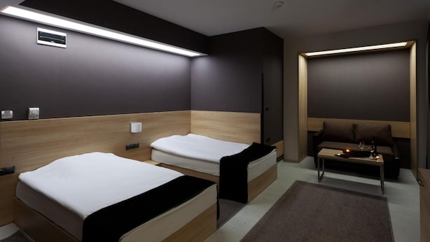 A modern hotel room with two beds, a sofa, a small table with a candle, and a wall-mounted TV. - Hotel Idol (Targovishte)
