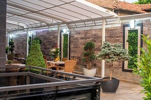 A covered outdoor seating area with tables and chairs, potted plants, and a stone wall with greenery. - Hotel Idol (Targovishte)