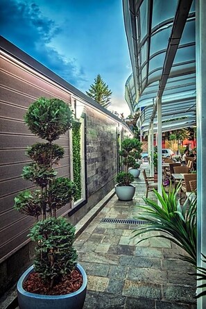 A narrow outdoor corridor with potted plants, stone flooring, and a modern building with large windows. - Hotel Idol (Targovishte)