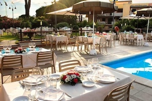 An outdoor dining area with tables set for a meal, a pool, and umbrellas providing shade. - Villagio La Darsena (Scarlino)