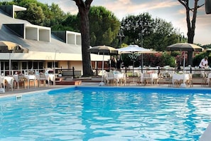 A swimming pool with tables and umbrellas, surrounded by trees and a building with a sloped roof. - Villagio La Darsena (Scarlino)