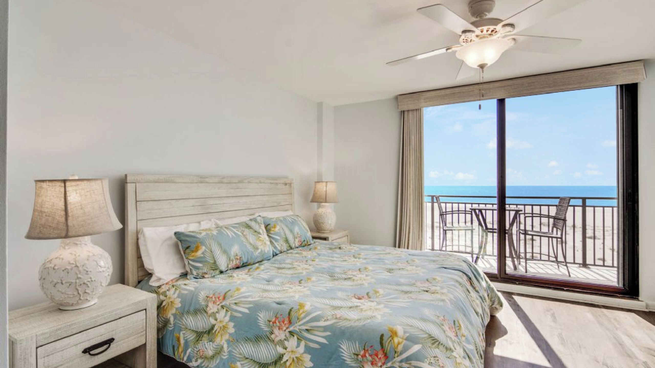 A bedroom with a bed, two bedside lamps, a nightstand, and a view of the beach through large windows.