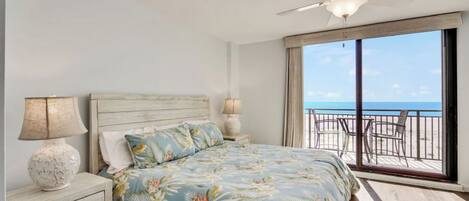 A bedroom with a bed, two bedside lamps, a nightstand, and a view of the beach through large windows.