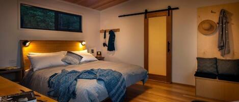 Ein gemütliches Schlafzimmer mit einem hölzernen Kopfteil, einem Bett mit blauer Decke, einem Nachttisch, einem Holztisch und einem Kleiderschrank, an dessen Wand ein Hut hängt.