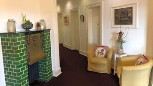 A hallway with a green-tiled fireplace, a yellow armchair with a red and white pillow, and a painting on the wall. - Fresena (Hamburg)