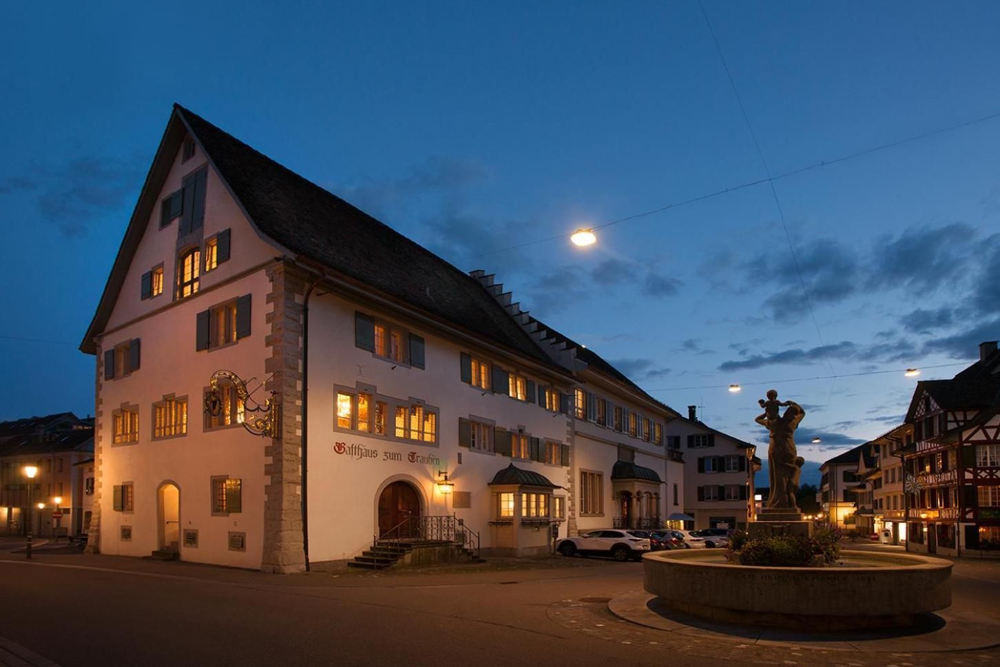 A traditional building with a sign 'Saalhaus zum Trauben' at the entrance, a staircase leading to the main door, and a statue in the foreground.