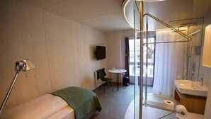A hotel room with a bed, a desk, a chair, a TV, a bathroom with a sink and mirror, and a window with curtains. - Gasthaus Zum Trauben (Weinfelden)