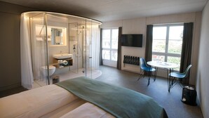 A modern hotel room with a bed, a glass-enclosed shower, a sink, a small table with chairs, a TV, and large windows. - Gasthaus Zum Trauben (Weinfelden)