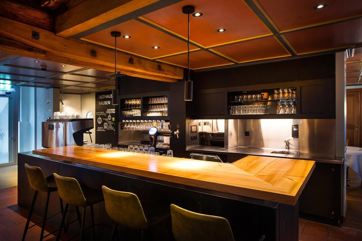 Eine Bar mit einer langen Holztheke, Barhocker und einer gut beleuchteten Decke mit Einbaustrahlern.