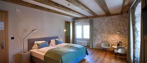 Une chambre avec un plafond en bois, un mur en pierre, un lit recouvert d’une couverture verte, des lampes de chevet, une petite table et une chaise.