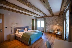 A bedroom with a wooden ceiling, stone wall, a bed with a green blanket, bedside lamps, a small table, and a chair. - Gasthaus Zum Trauben (Weinfelden)