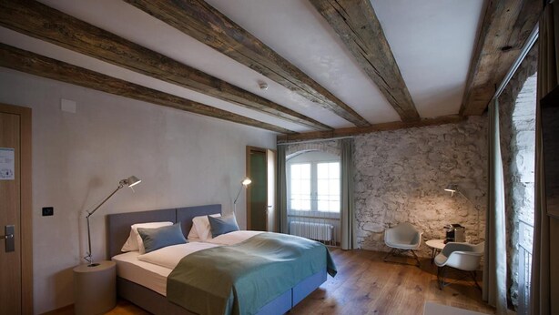 A bedroom with a wooden floor, a bed with a green blanket, a desk with a chair, and a stone wall. - Gasthaus Zum Trauben (Weinfelden)