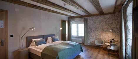 A bedroom with a wooden floor, a bed with a green blanket, a desk with a chair, and a stone wall.