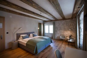 A bedroom with a wooden floor, a bed with a green blanket, a desk with a chair, and a stone wall. - Gasthaus Zum Trauben (Weinfelden)
