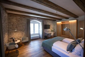 Un dormitorio con pared de piedra, techo de madera, cama, escritorio, silla, televisor y lámpara de noche.