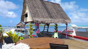 A wooden deck with a table and chairs, a thatched-roof structure, and a view of the ocean and boats.