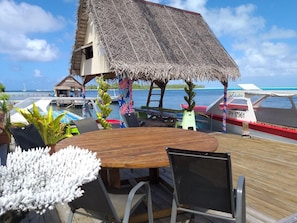 A wooden deck with a table and chairs, a thatched-roof structure, and a view of the ocean and boats.