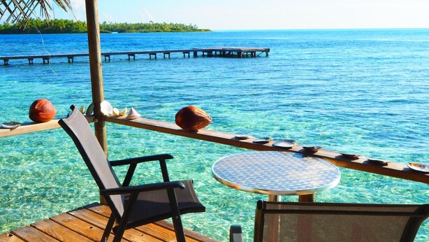 A wooden deck with a round table and chairs, overlooking clear blue water and a distant pier.