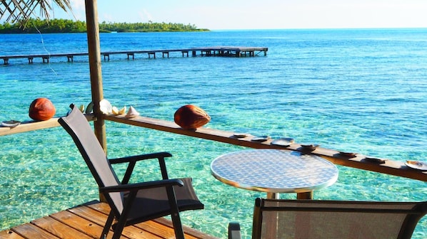 A wooden deck with a round table and chairs, overlooking clear blue water and a distant pier.