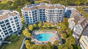 Un hotel de varios pisos con piscina central rodeada de sillones de descanso y palmeras.