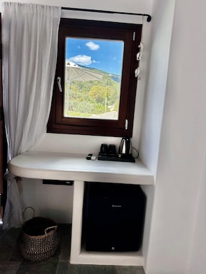 Ein Zimmer mit Fensterblick auf eine gebirgige Landschaft, ein weißer Schreibtisch mit Kaffeemaschine und ein schwarzes elektronisches Gerät auf einem Regal.