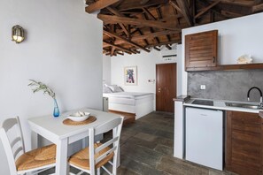 Uma cozinha compacta com máquina de lavar louça embutida, um espaço de refeições com mesa branca e cadeiras de vime, e um quarto com cama branca e um quadro na parede.