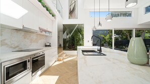 A modern kitchen with a large island, built-in oven, and a hanging chair. - Meli Beach House by Experience Jervis Bay (Callala Beach)