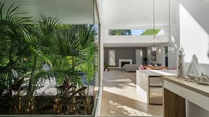 A modern interior with a kitchen, dining area, and living space, featuring a large window and indoor plants. - Meli Beach House by Experience Jervis Bay (Callala Beach)